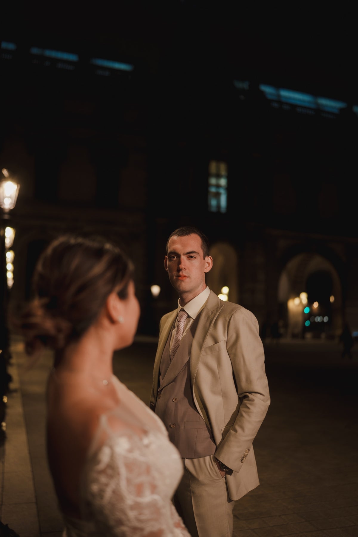 A man in a beige three-piece suit and a woman in a white lace wedding dress standing in a dimly lit nighttime outdoor setting.