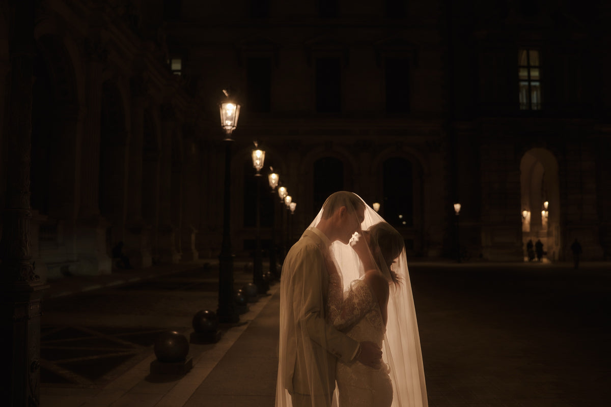 A bride in a white wedding gown and veil and a groom in a light-colored suit embrace and kiss in a dimly lit nighttime outdoor setting with warm street lamps and a grand building in the background.