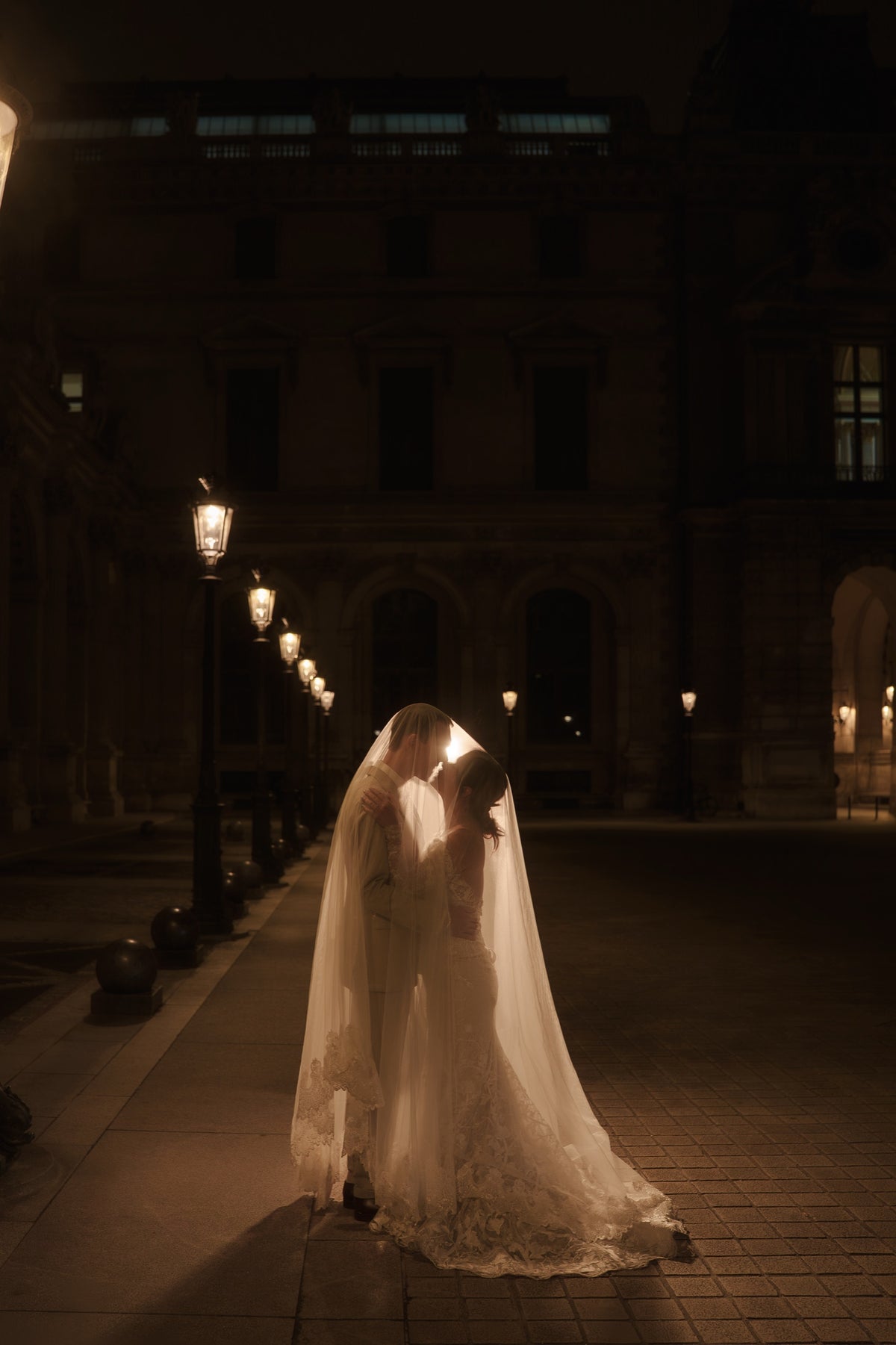 A bride in a long white lace wedding gown and veil embraces and kisses a man on a dark cobblestone street at night, with warm streetlights and a grand building in the background.