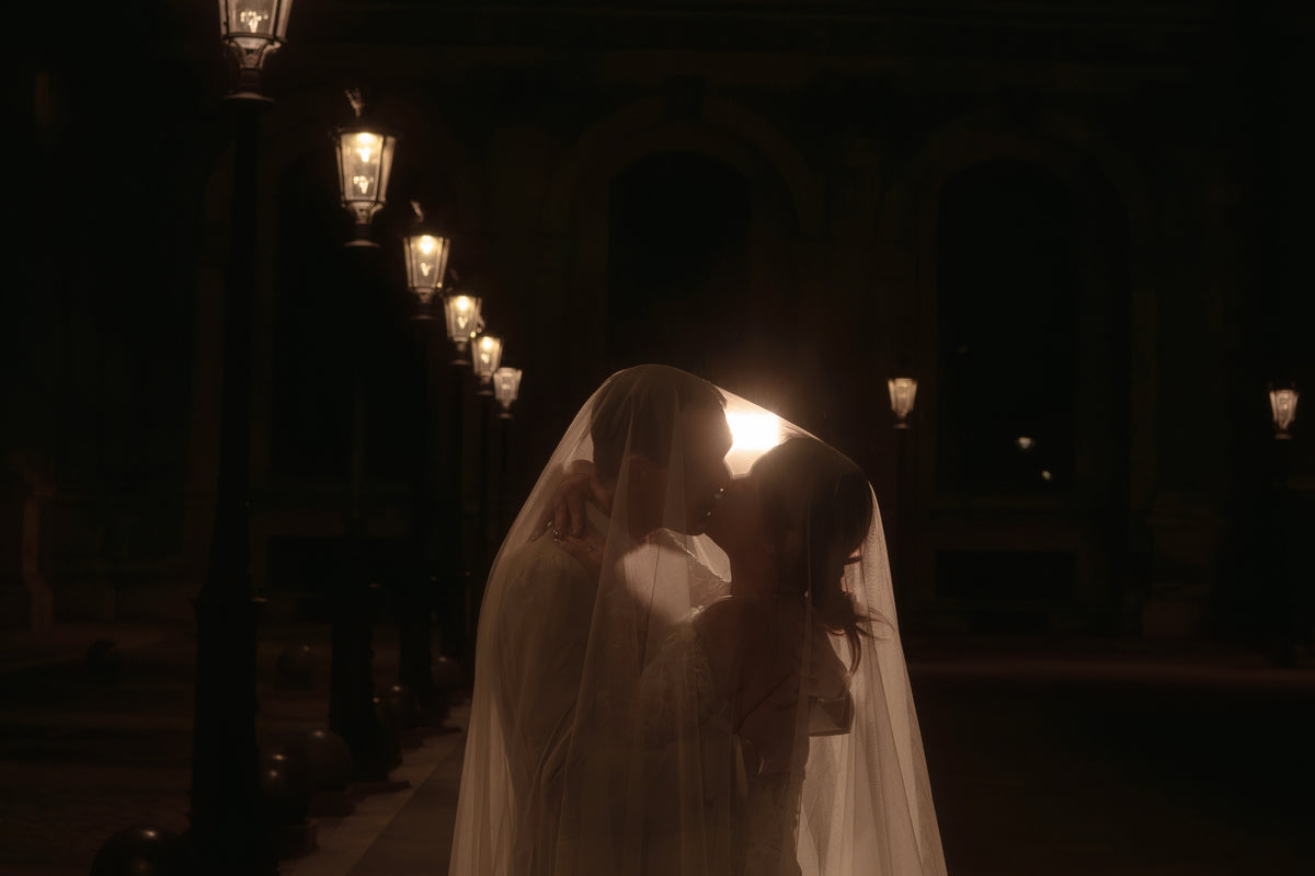 A bride in a white wedding gown and veil embracing and kissing her groom in a dimly lit outdoor setting at night, with warm street lamps lining the background.