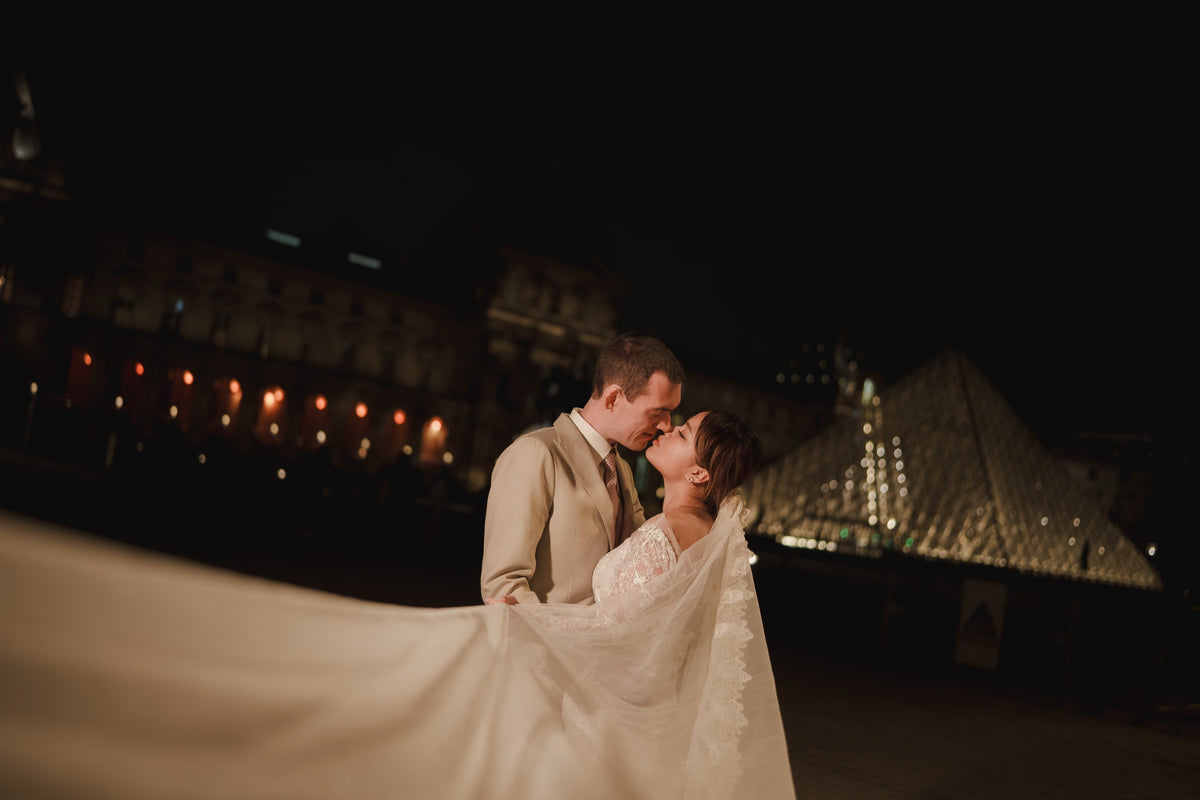 A bride in a white lace wedding gown with a long veil and a groom in a beige suit kiss under the lights of the Louvre Pyramid at night.
