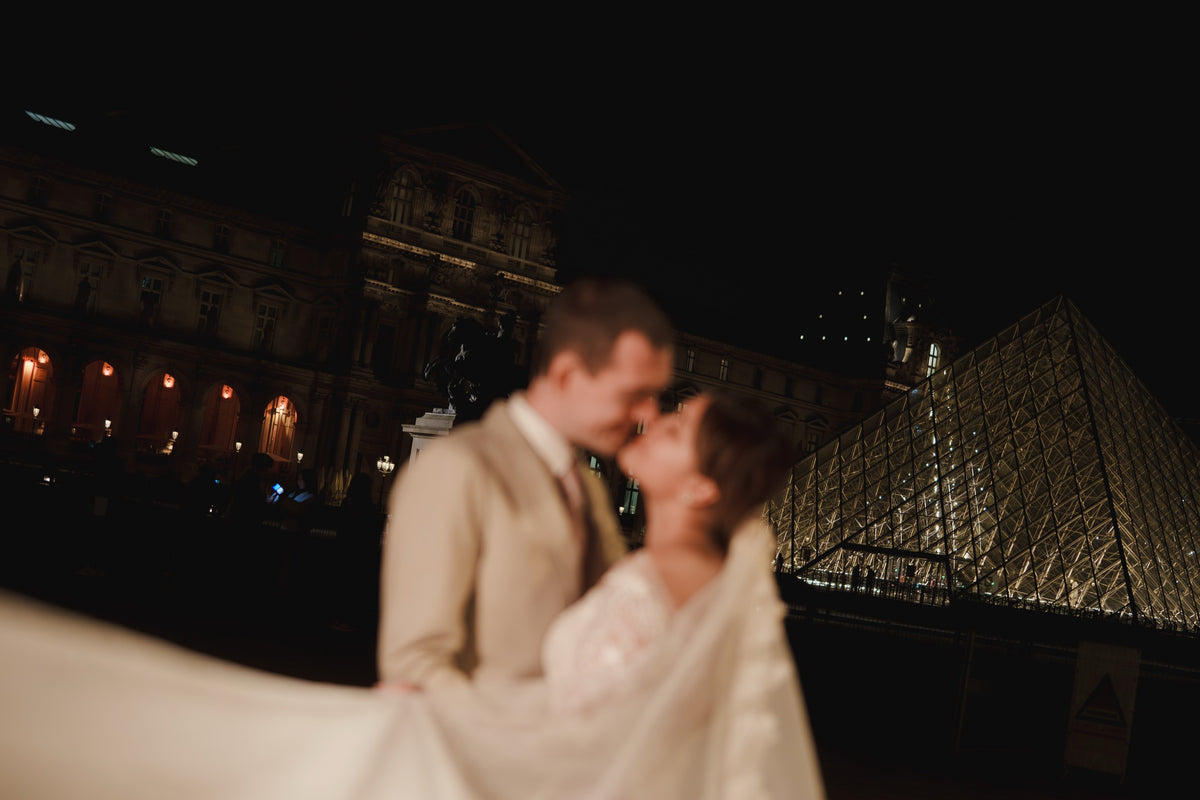A couple embracing and kissing at night in front of the Louvre Museum, with the bride's white veil flowing across the foreground and the illuminated glass pyramid and classical building in the background.