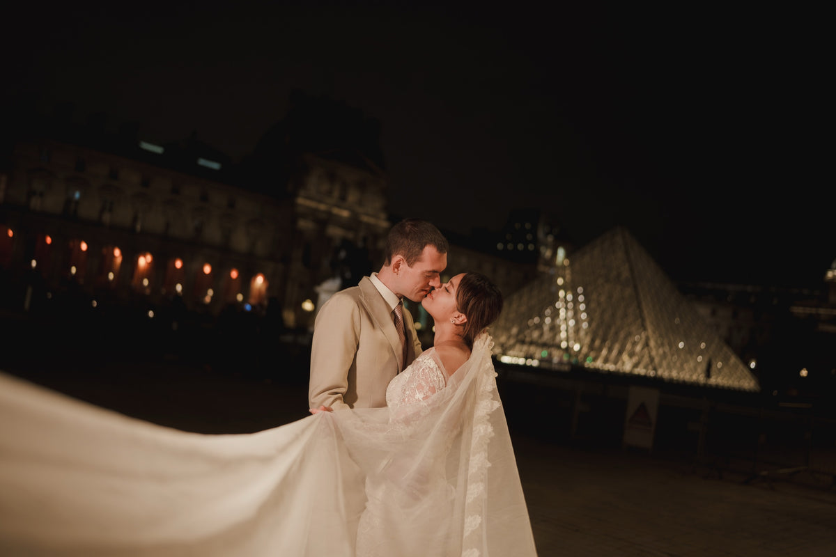 A bride in a white lace wedding gown with a long sheer veil and a groom in a beige suit kiss in front of the illuminated Louvre Pyramid at night.