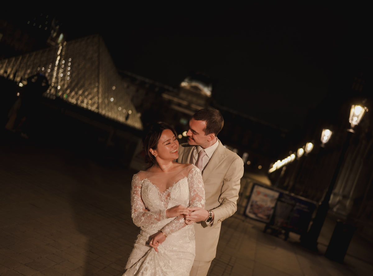 A bride in a white lace wedding gown and a groom in a beige suit embrace at night in front of the illuminated Parisian Pyramid.