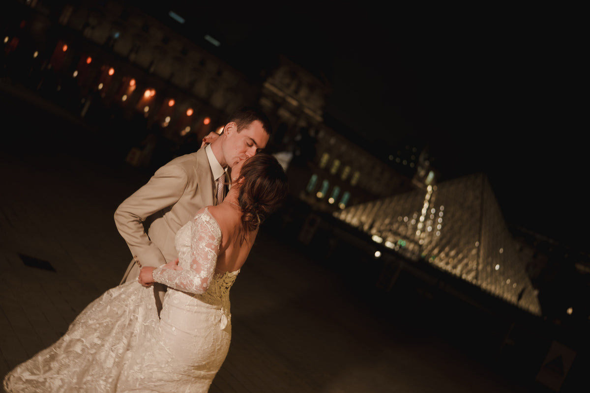 A bride in a cream lace wedding gown and a groom in a beige suit embrace and kiss outdoors at night, with a softly blurred illuminated building and glass pyramid in the background.
