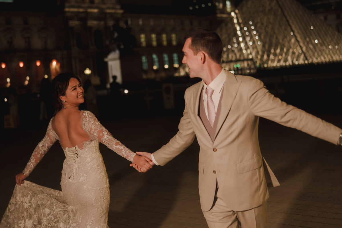 A bride in a fitted lace wedding gown with a backless design and a man in a light beige suit and pink tie holding hands while dancing at night in front of a city building with illuminated glass structures.