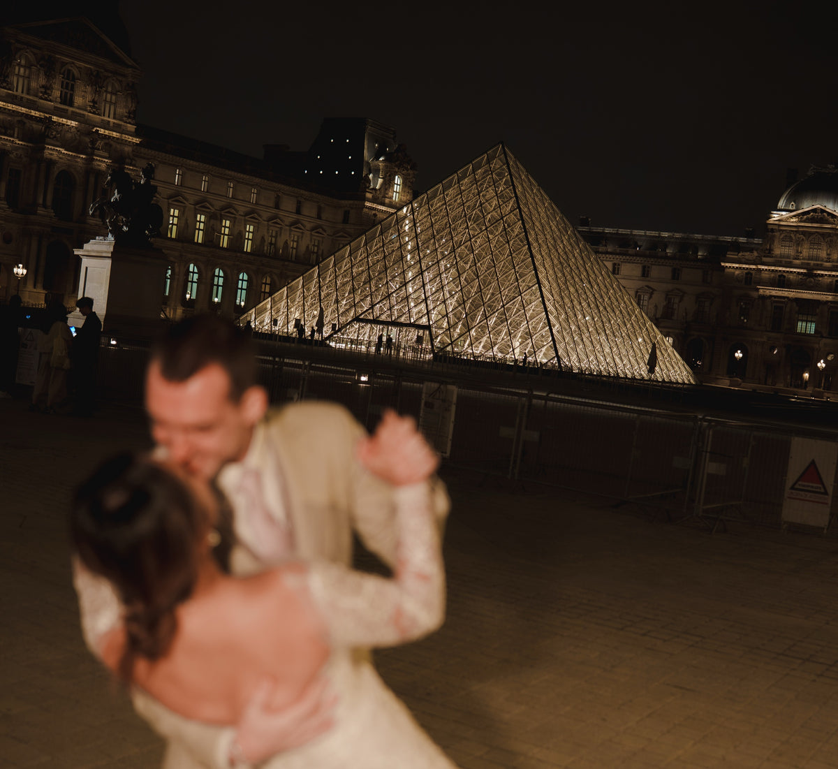 A couple embracing and dancing in front of the illuminated Louvre Pyramid at night, with the Parisian Opera House in the background.