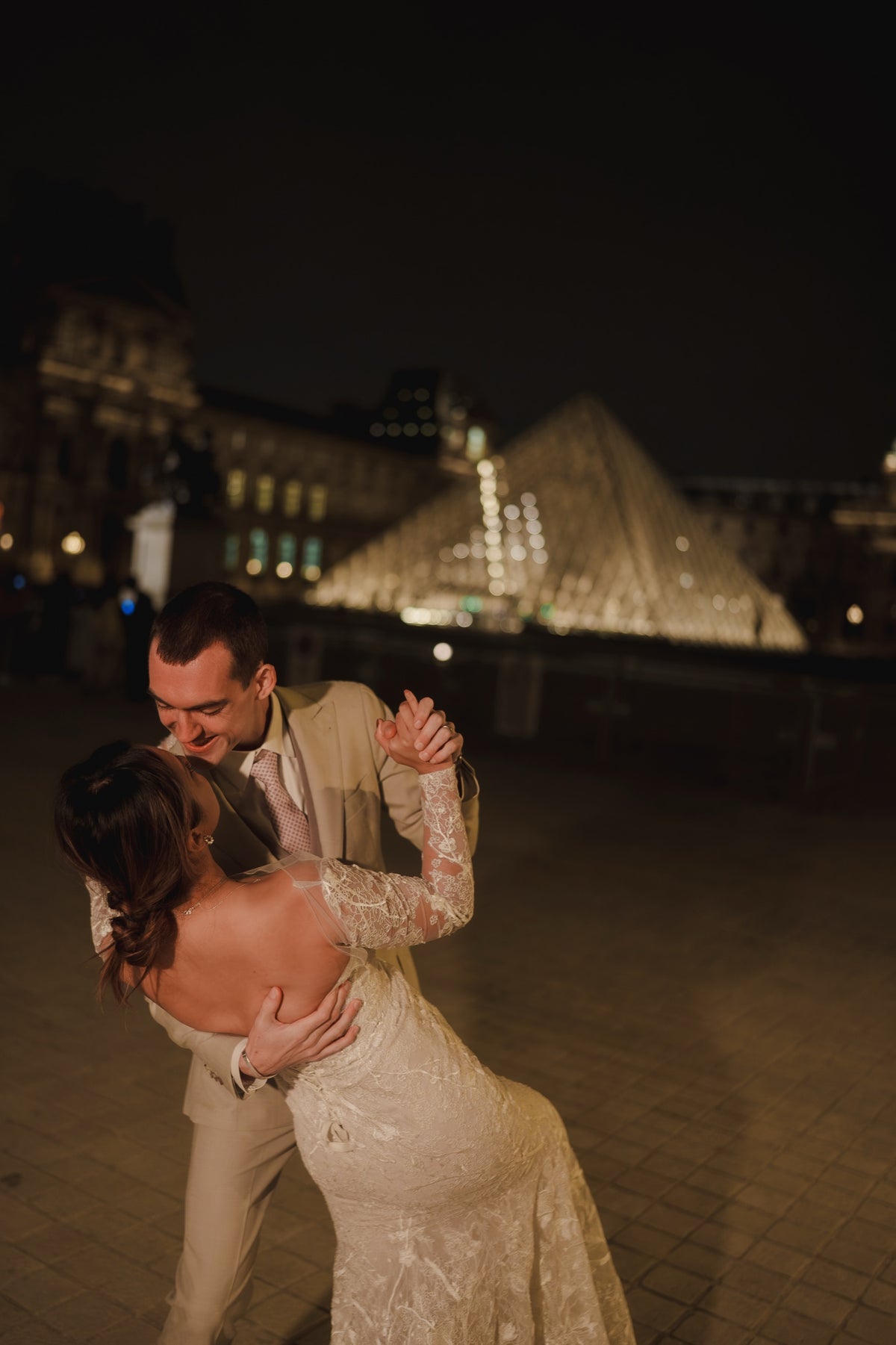 A bride in a long-sleeved lace wedding gown and a groom in a light beige suit embrace and dance in front of the illuminated Louvre Pyramid at night.