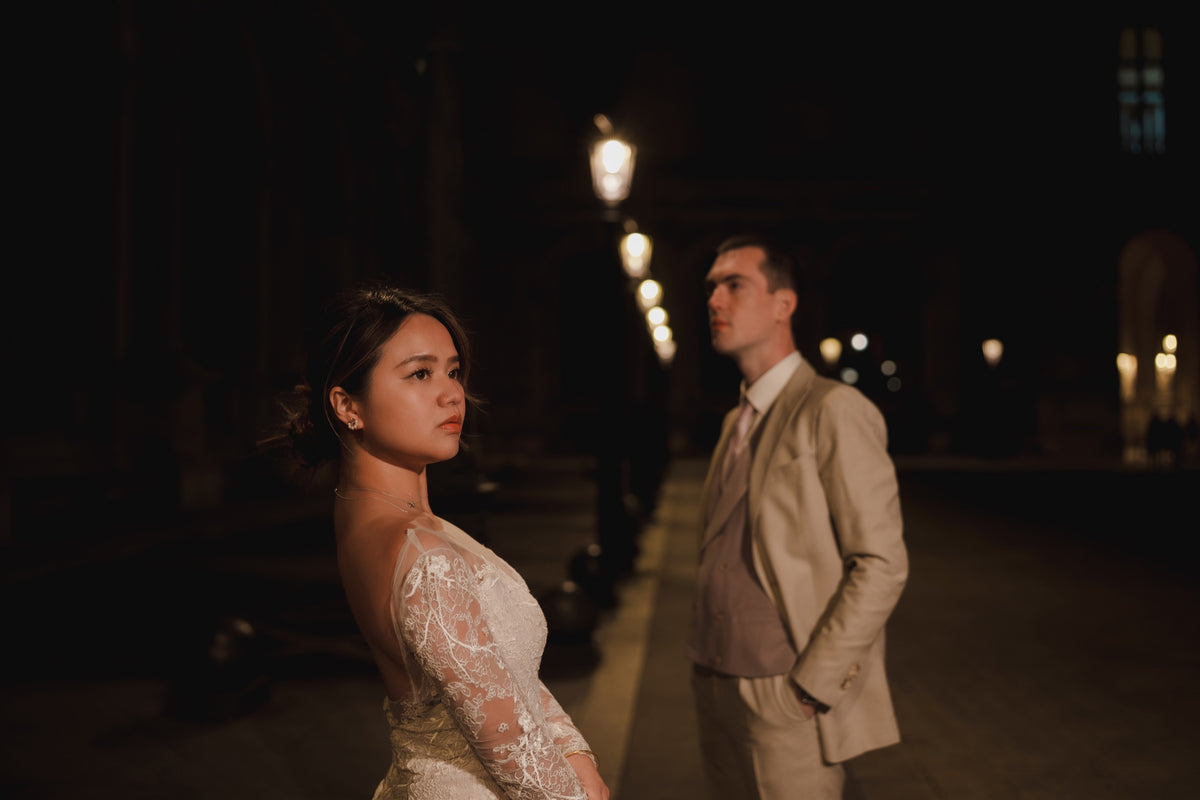 A bride in a lace wedding gown and a groom in a beige suit standing on a dark street at night with warm streetlights in the background.