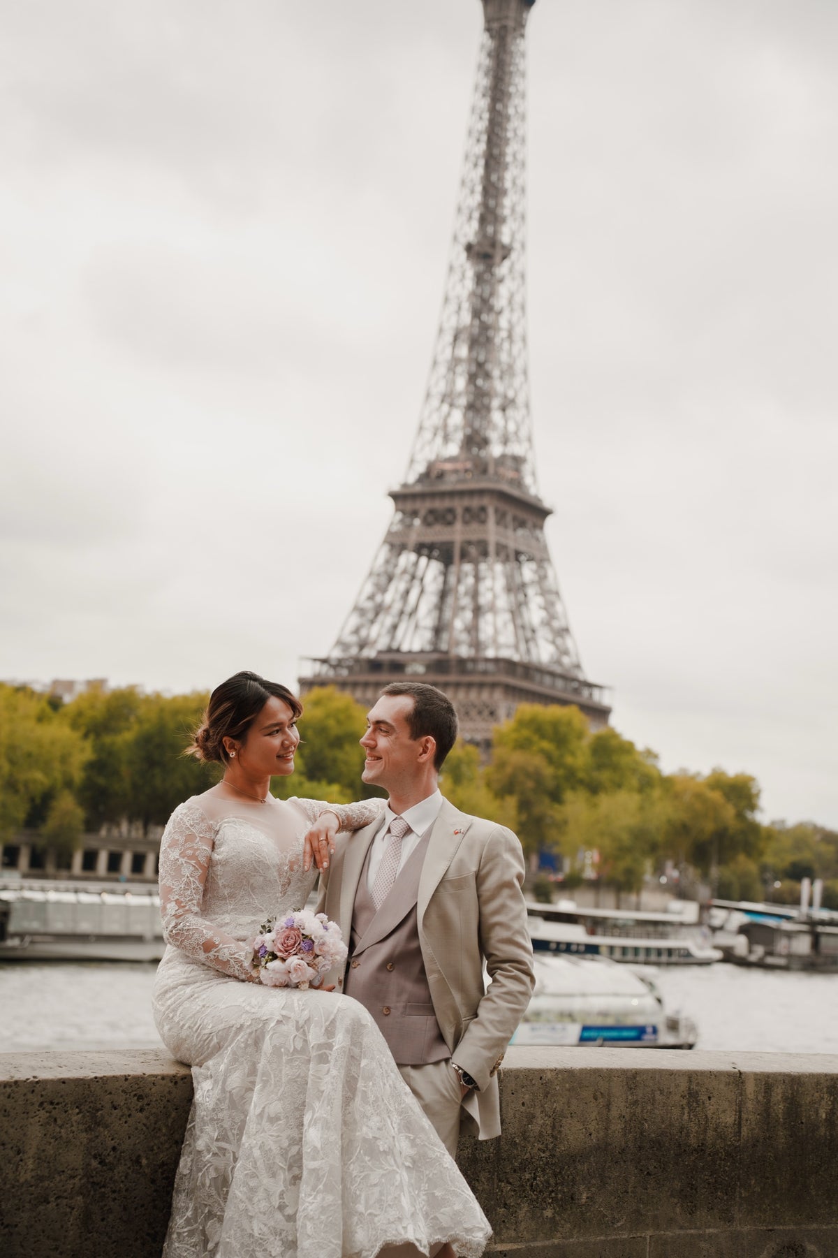 A bride in a white lace wedding gown and a groom in a light beige suit sit together on a stone ledge by the Seine River, with the Eiffel Tower and boats in the background under an overcast sky.