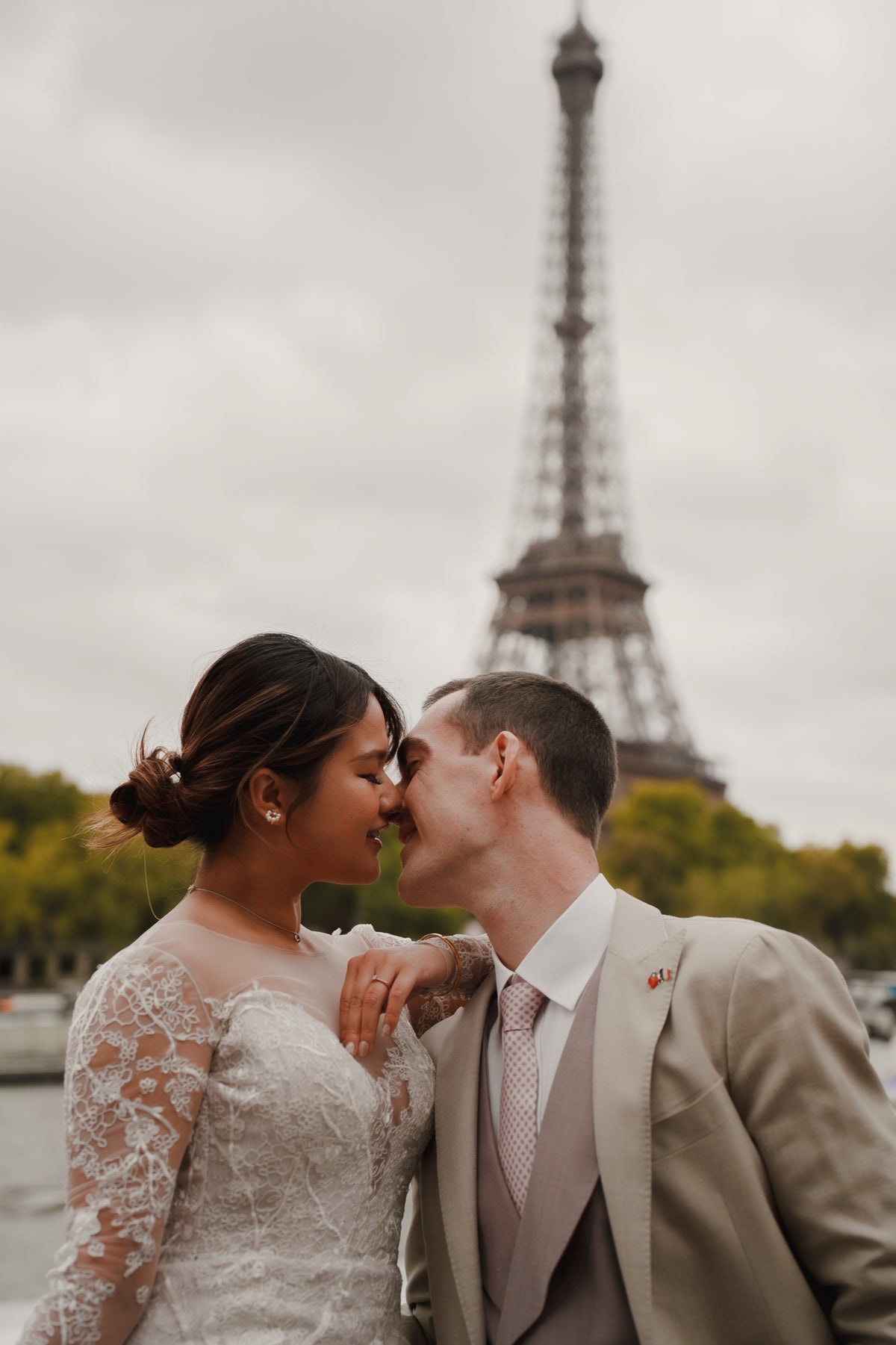 A bride in a white lace wedding gown and a groom in a beige suit kiss in front of the Eiffel Tower under an overcast sky.