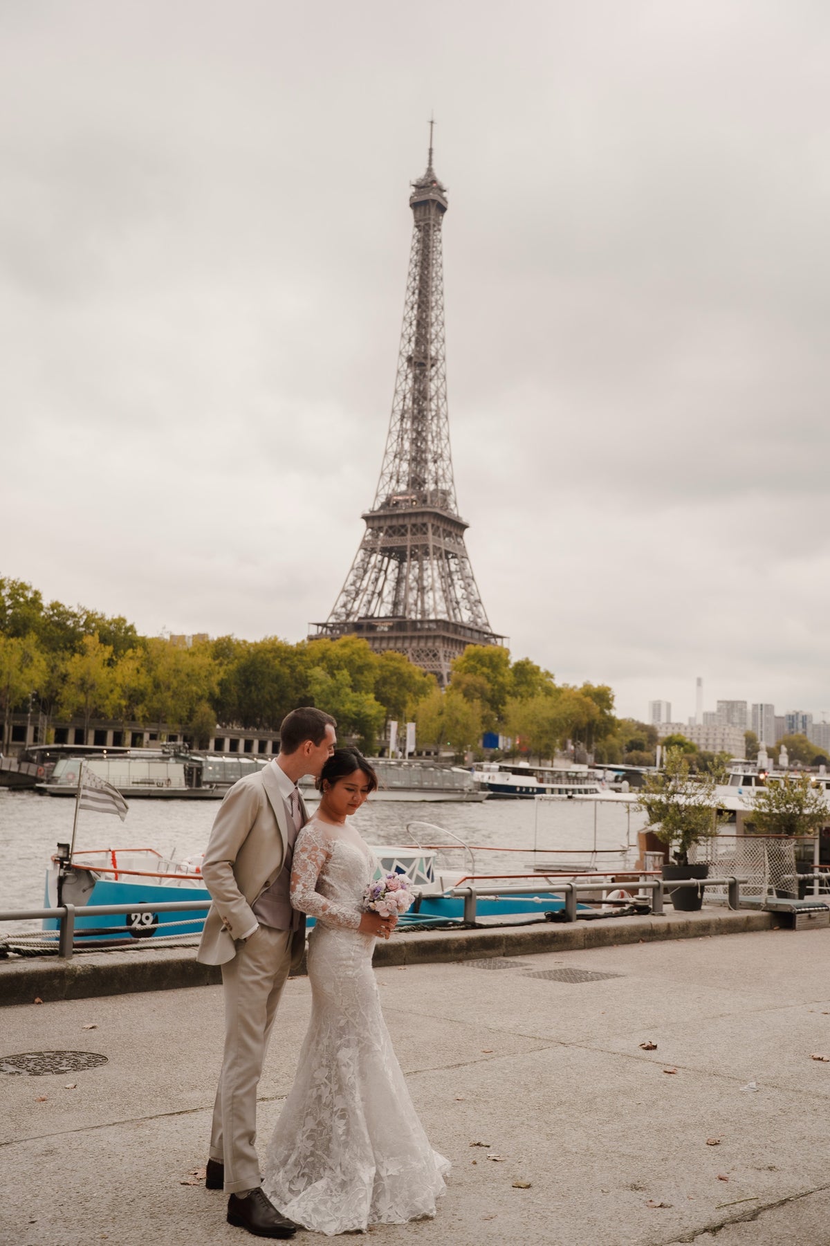 A bride in a white lace wedding gown and a groom in a beige suit stand on a riverside promenade with the Eiffel Tower in the background under an overcast sky.