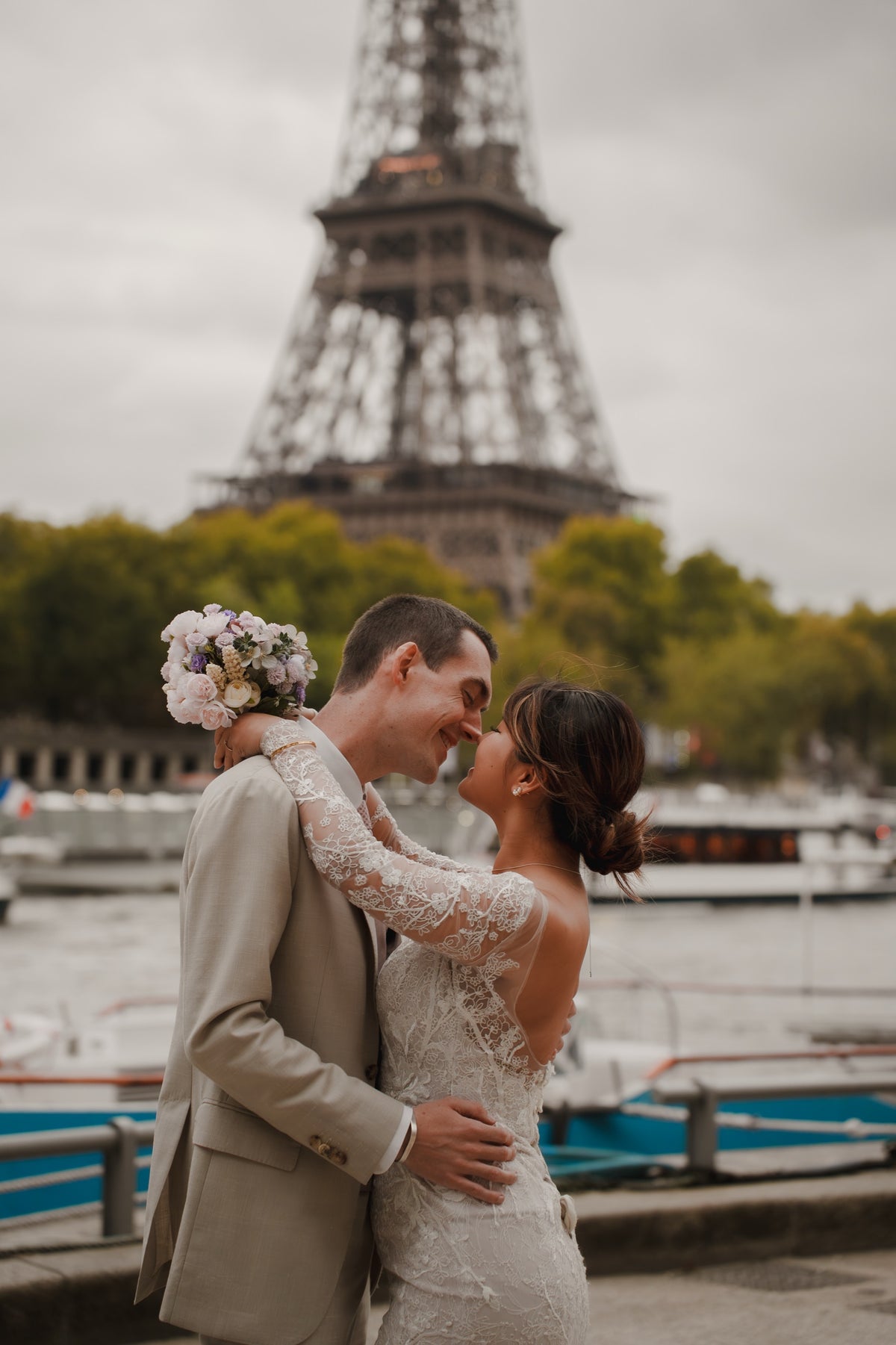 A bride in a white lace wedding gown and a groom in a beige suit embrace in front of the Eiffel Tower, with boats and green trees in the background.