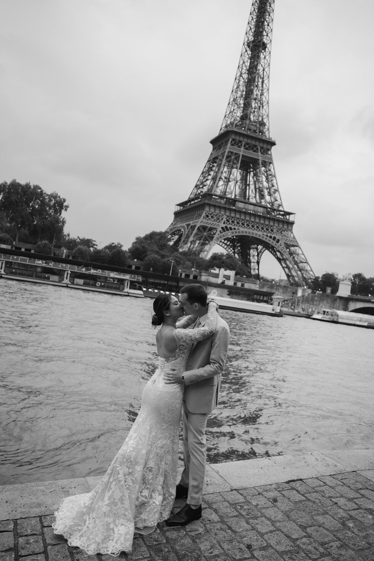 A black and white photograph of a bride and groom embracing on a riverside promenade with the Eiffel Tower in the background.