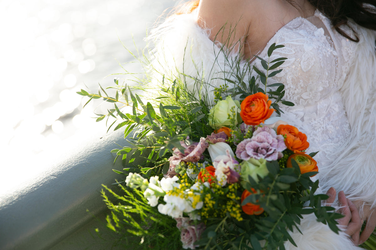 A close-up of a bride in a white lace wedding gown holding a vibrant floral bouquet with orange roses, purple lisianthus, white flowers, and lush green foliage, set against a bright, sunlit outdoor background.