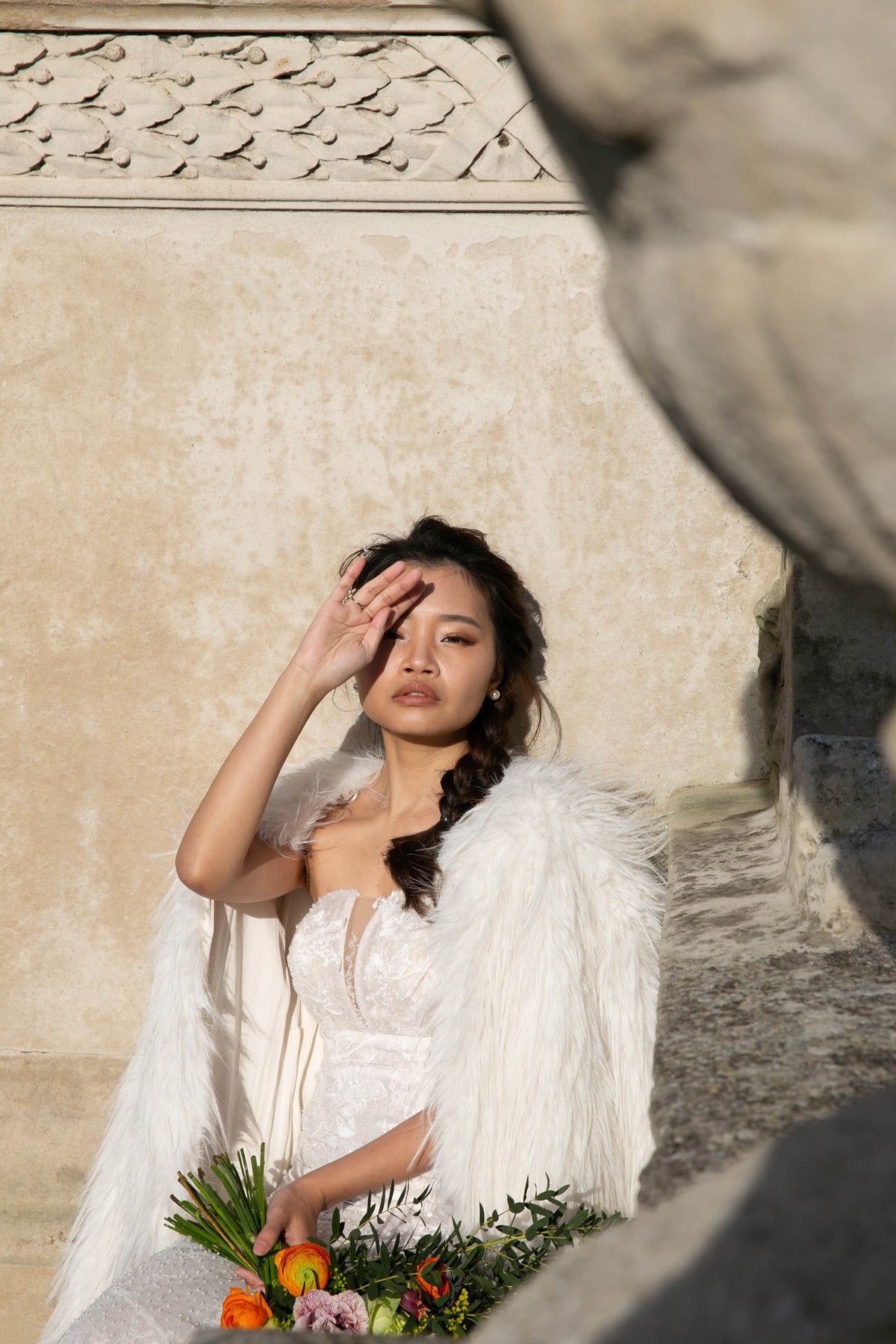 A woman in a white lace bridal gown with a fluffy white feathered shawl sits outdoors against a textured beige wall, holding a bouquet of flowers.