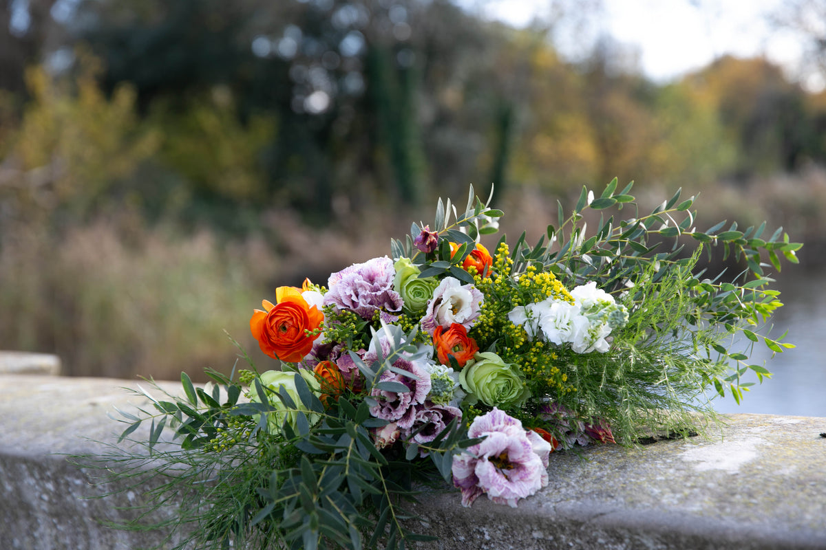 A rustic wildflower bouquet with orange, purple, white, and green flowers resting on a weathered stone bridge with a blurred outdoor background.