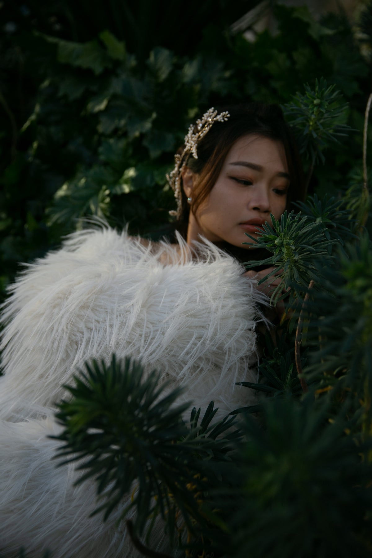 A woman wearing a voluminous white faux-fur garment or wrap and a delicate gold headpiece, posed among dark green foliage in a moody outdoor setting.
