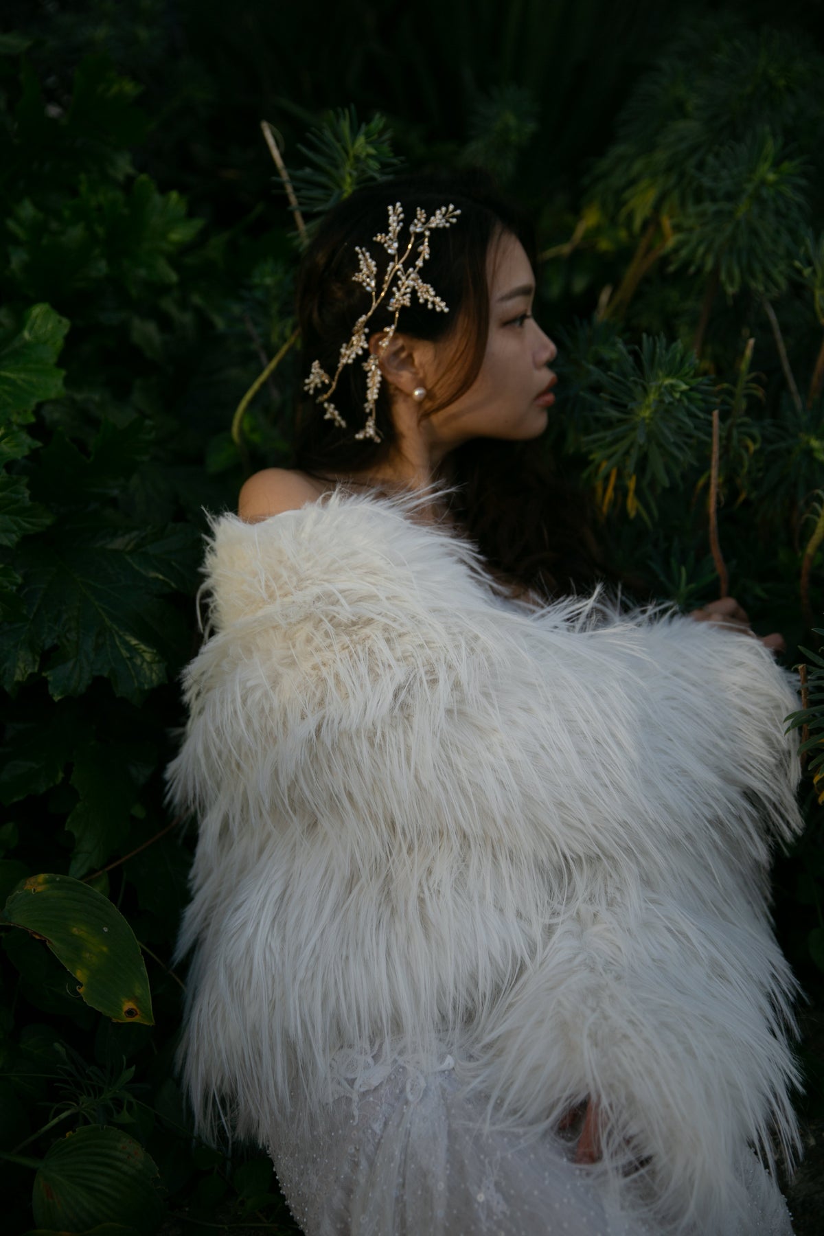 A woman in profile wearing a voluminous white faux-fur shawl and a matching white dress, styled with a gold leaf headpiece, set against a dark green foliage background.