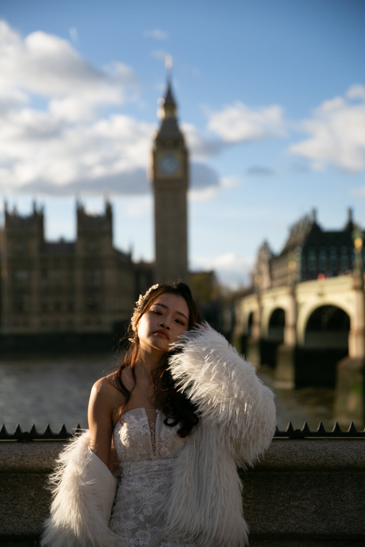 A woman in a white lace dress and fluffy white faux-fur stole stands outdoors in front of a blurred London backdrop featuring Big Ben and the Thames Bridge.