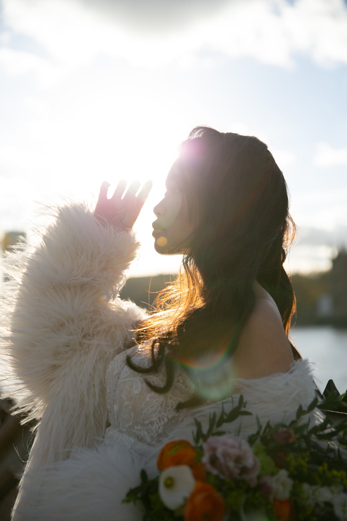A woman in a white off-the-shoulder lace dress and fluffy white faux-fur jacket stands outdoors in bright backlit sunlight, holding a bouquet of orange, pink, and white flowers.