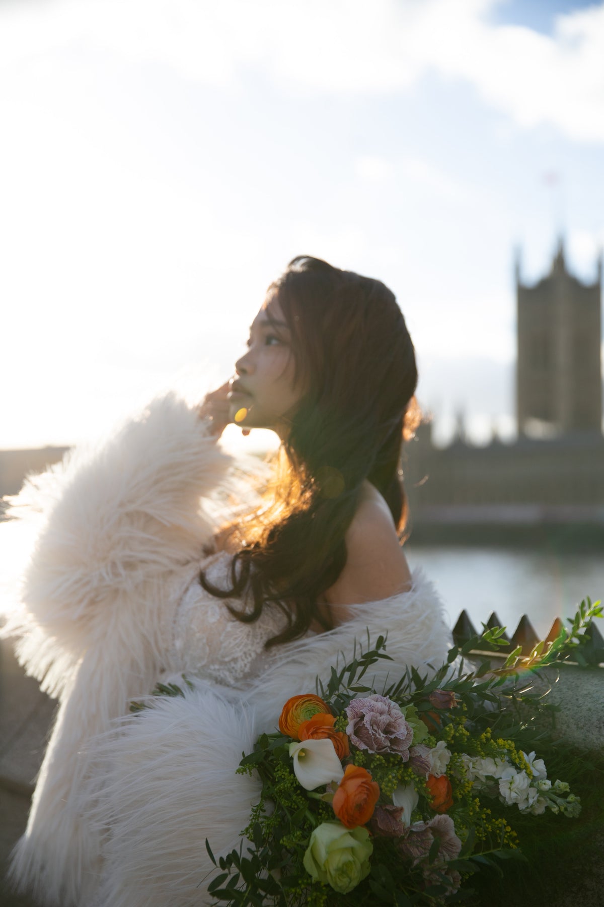 A woman in a voluminous white faux-fur coat and white dress stands outdoors in warm backlighting, holding a colorful bouquet of flowers. The background is a softly blurred outdoor scene with water and a tower.