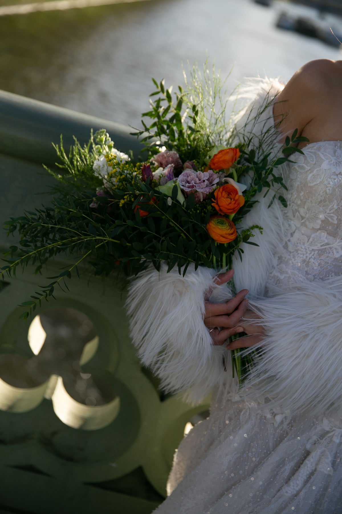 A close-up lifestyle shot of a bride in a white lace wedding gown holding a vibrant floral bouquet with orange ranunculus, pink flowers, and green foliage, while wearing a white feather-trimmed wrap on a bridge.