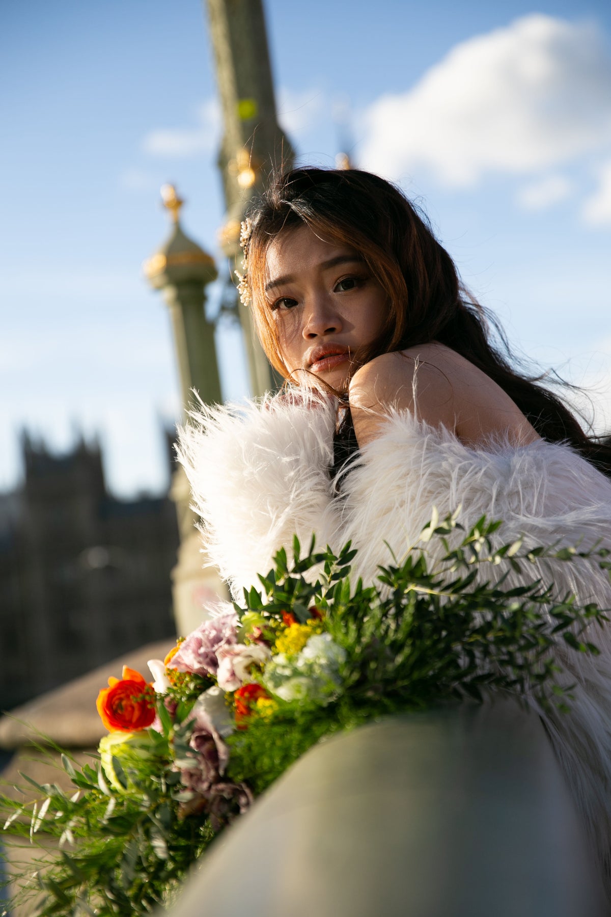 A woman outdoors wearing a voluminous white feathered coat or dress, holding a colorful bouquet of flowers. The background shows a blurred outdoor scene with blue sky, clouds, and architectural elements.
