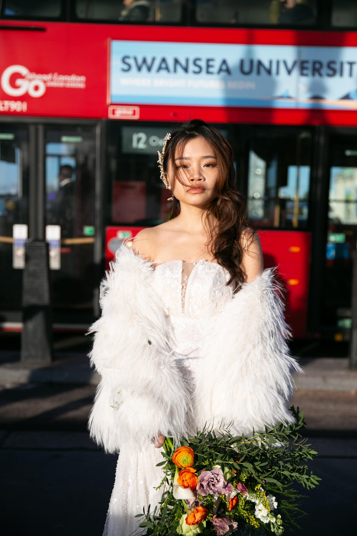 A woman wearing a white off-the-shoulder feathered dress and holding a floral bouquet stands in front of a red Swansea University bus.
