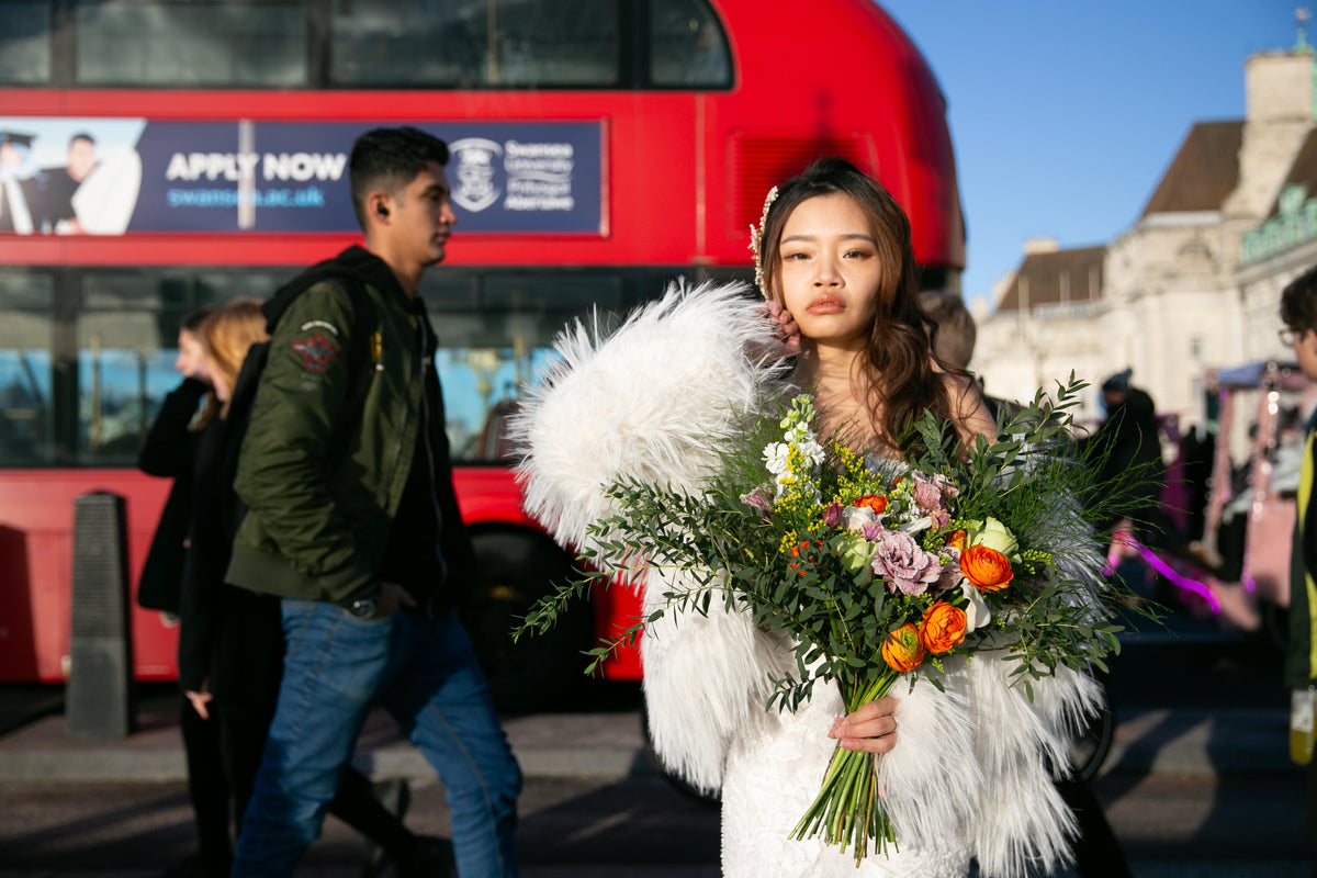 A woman in a white feathered bridal gown holds a large, vibrant bouquet of flowers on a busy city street with a red double-decker bus in the background.