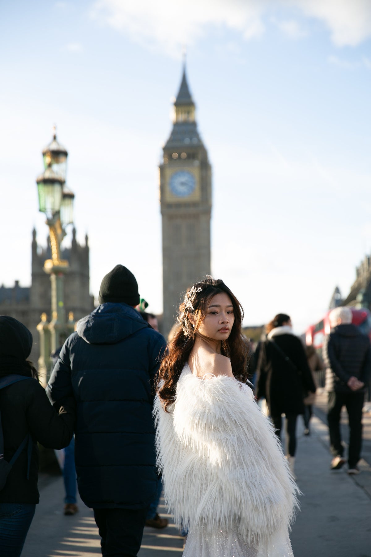 A woman wearing a voluminous white faux-fur wrap or dress stands on a bridge in London, with Big Ben in the background. Other pedestrians are visible in the foreground and around her.