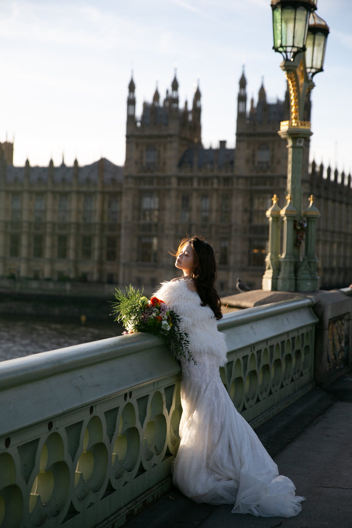 A woman in a white wedding gown and fluffy white shawl leans against the Westminster Bridge railing in London, holding a bouquet of flowers, with the Houses of Parliament and a lamppost in the background.