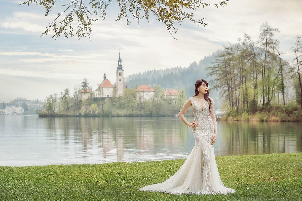 A woman wearing a sleeveless white lace mermaid wedding gown stands on a grassy lakeside with a scenic lake, church, and forested hills in the background.