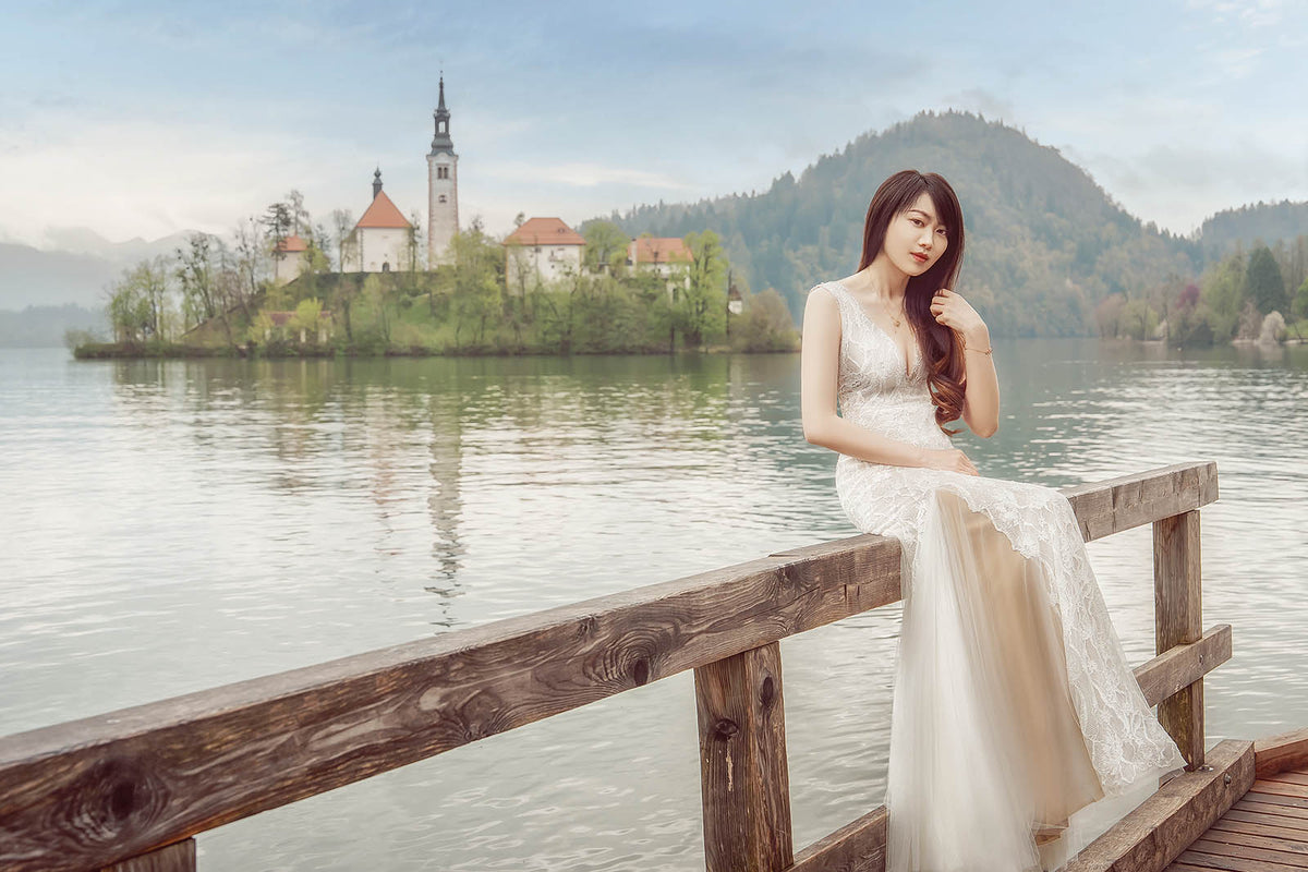 A woman in a sleeveless lace wedding gown sits on a wooden railing by a lake, with a small island church and forested mountains in the background.