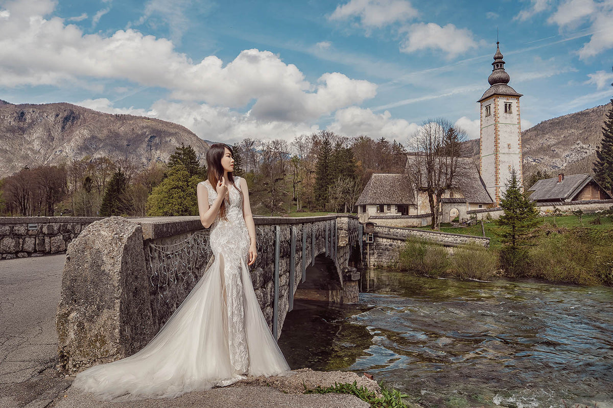 A woman in a sleeveless white lace wedding gown stands on a stone bridge beside a flowing river, with mountains, a church tower, and cloudy blue sky in the background.