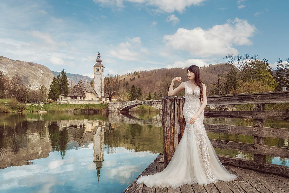 A woman in a sleeveless white lace wedding gown stands on a wooden dock beside a calm lake, with a church, mountains, and blue sky reflected in the water in the background.