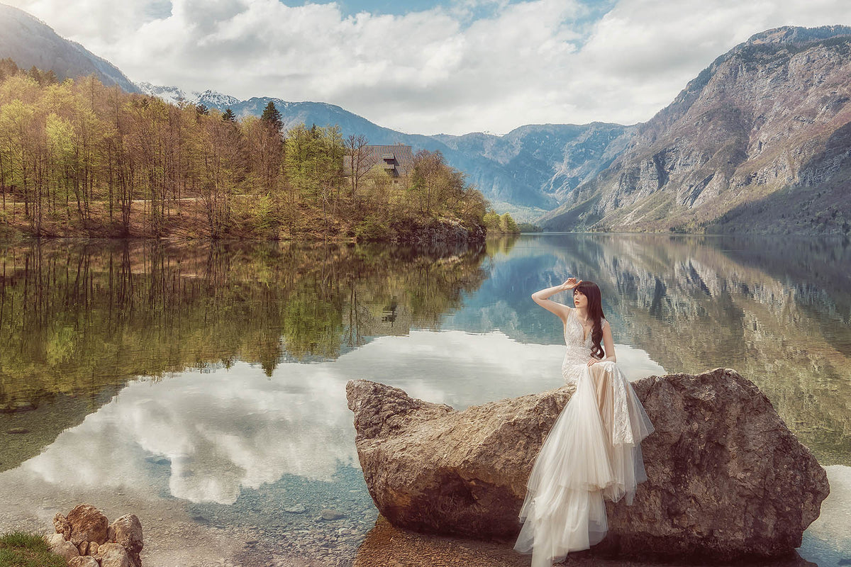 A woman in a flowing white wedding gown stands on a large rock by a calm lake, with mountains, trees, and cloudy reflections in the water behind her.