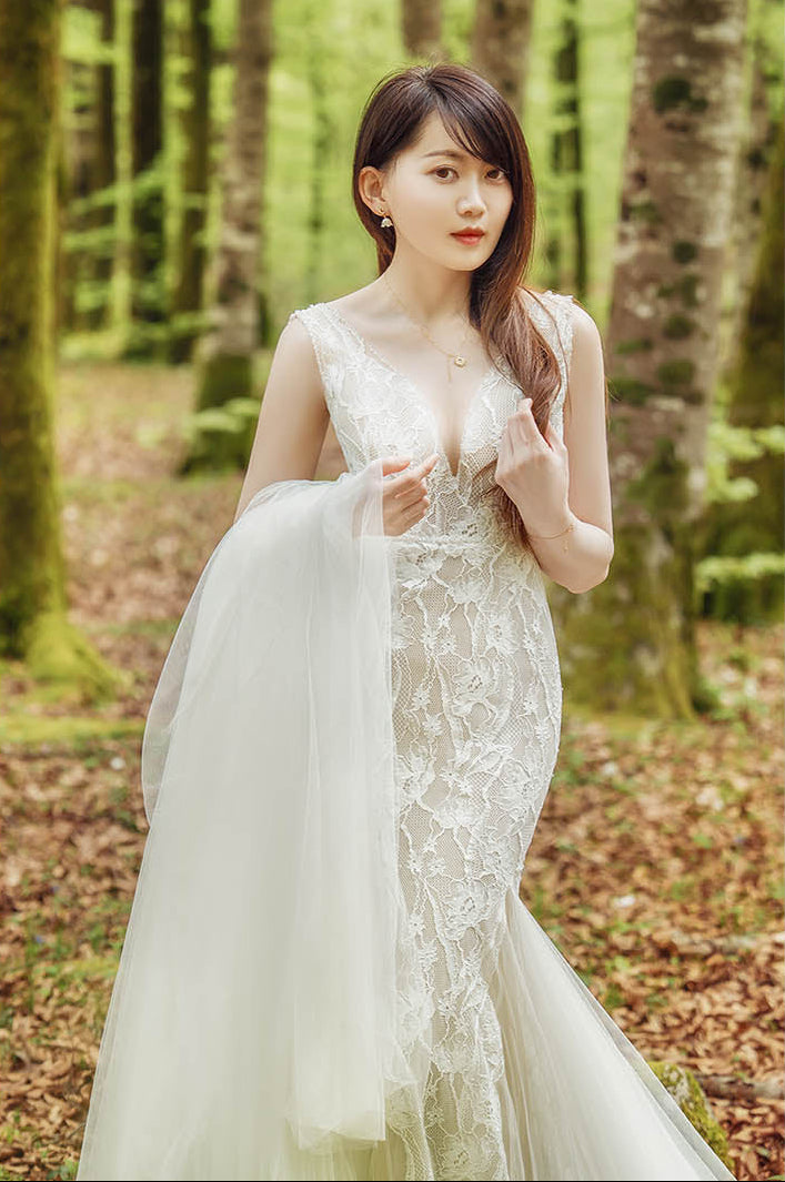 A woman wearing a sleeveless lace wedding gown stands in a forest with trees and fallen leaves visible in the background.