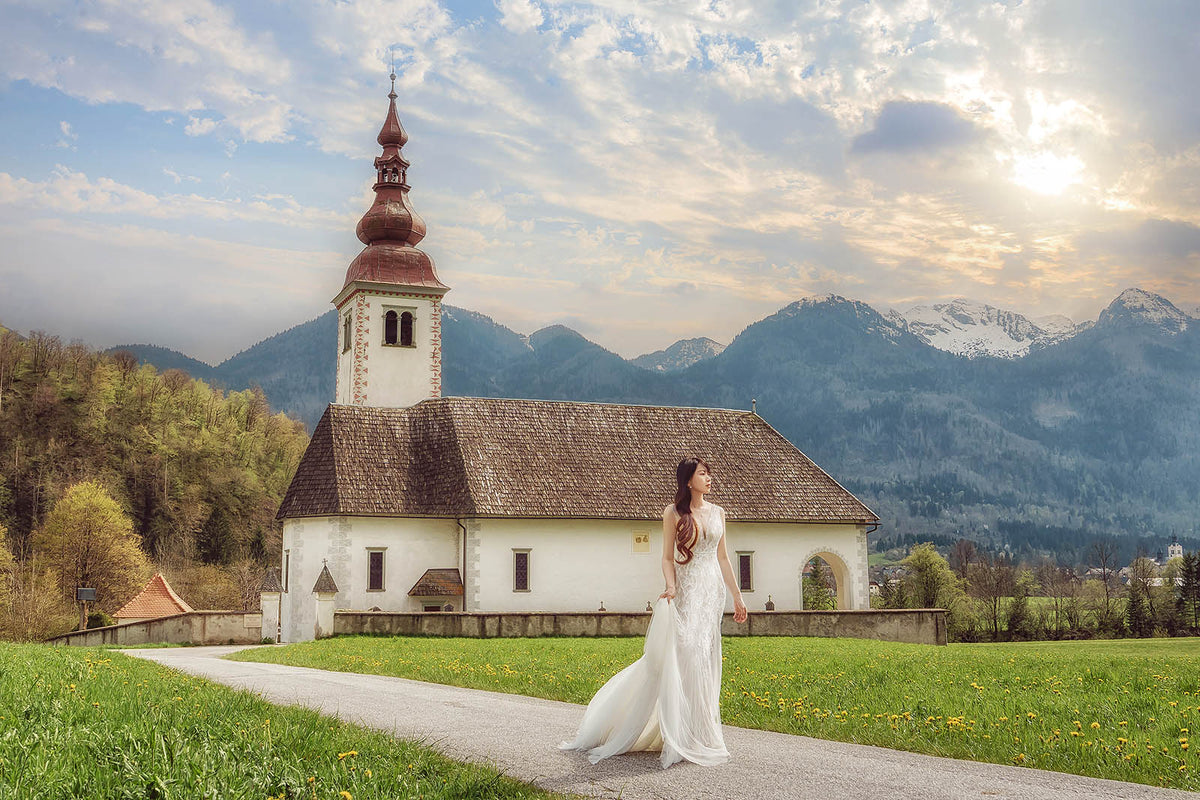 A woman in a white wedding gown stands on a path in front of a white church with a brown spire, surrounded by green grass, trees, and mountains under a cloudy sky with sunlight breaking through.