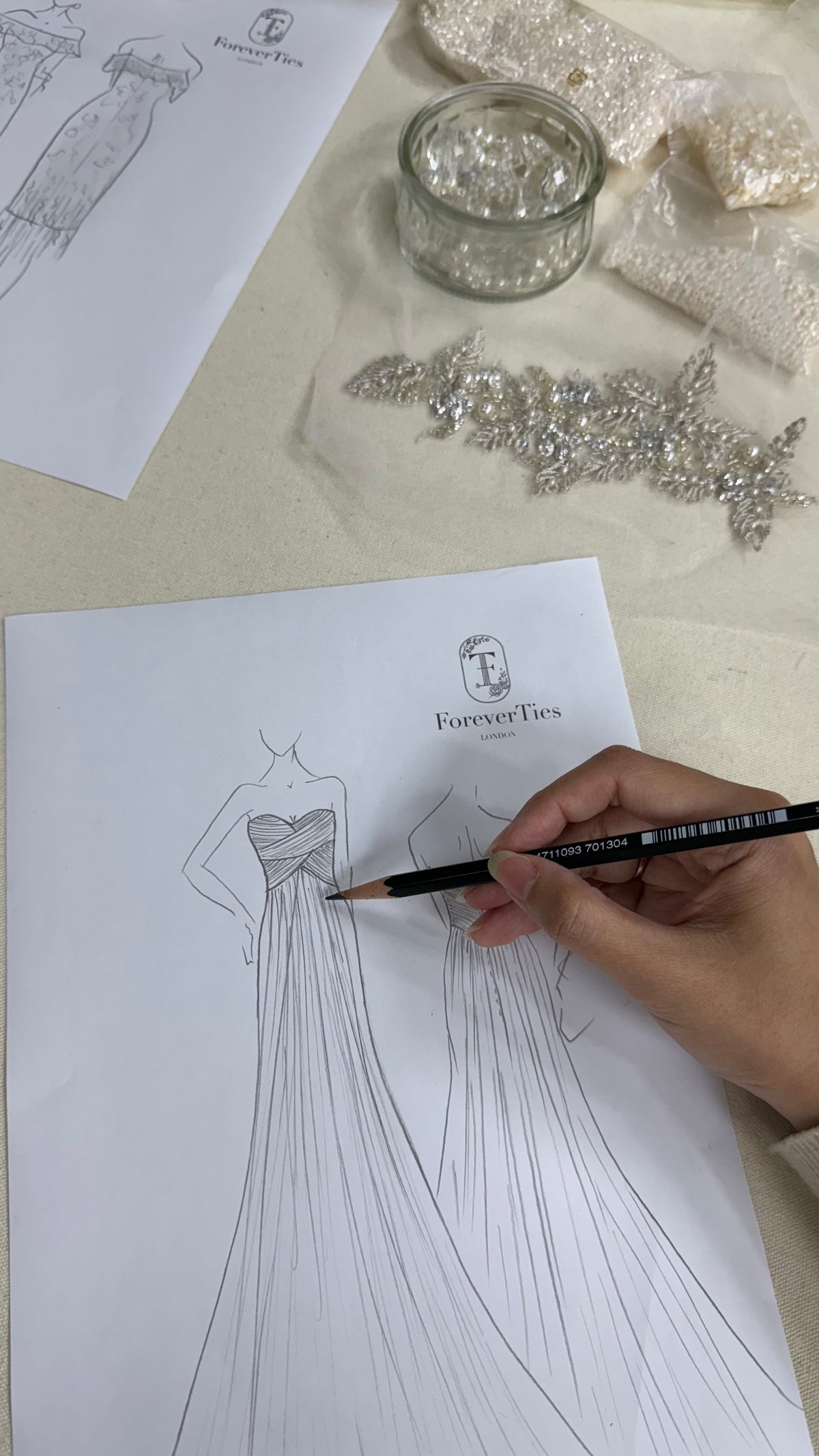 A bridal designer sketching a flowing wedding gown by hand, with crystal embellishments and design materials arranged nearby.